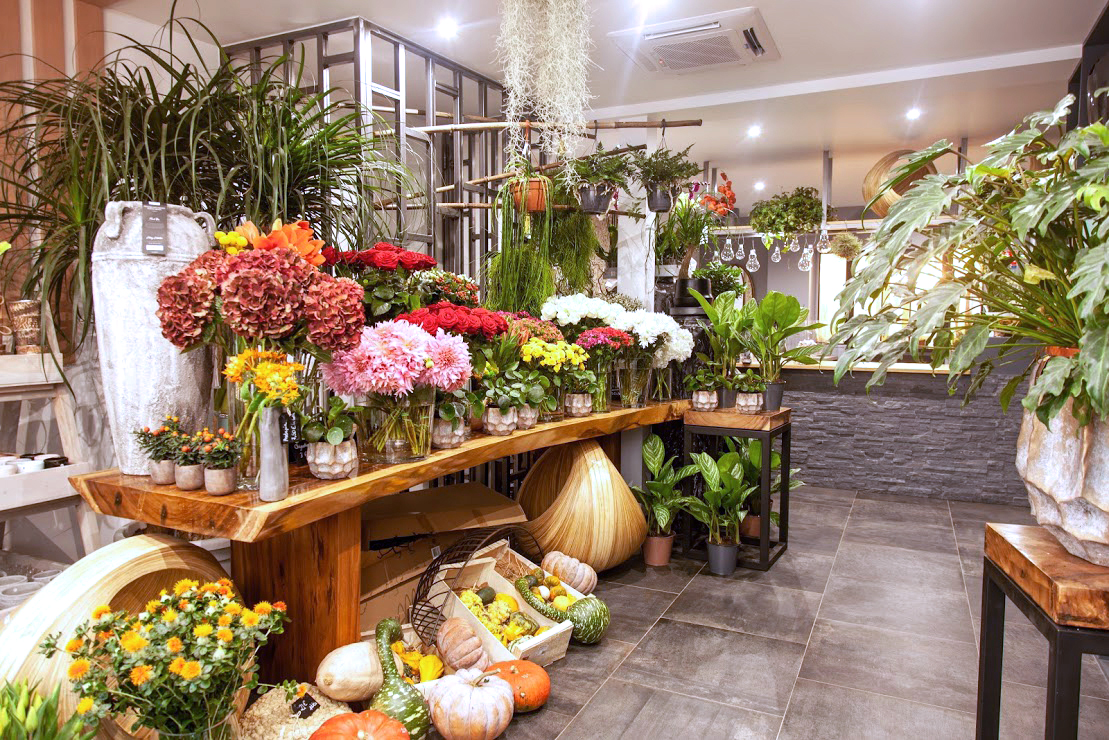 l’intérieur de la boutique : table en bois exotique sur la gauche, avec un assortiment de vases garnis de fleurs. Comptoir en face.